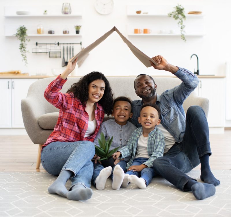 Multicultural family holding cardboard box above heads in form of house roof while sitting on carpet in kitchen. Smiling adults and children updating moving day checklist while resting on floor.