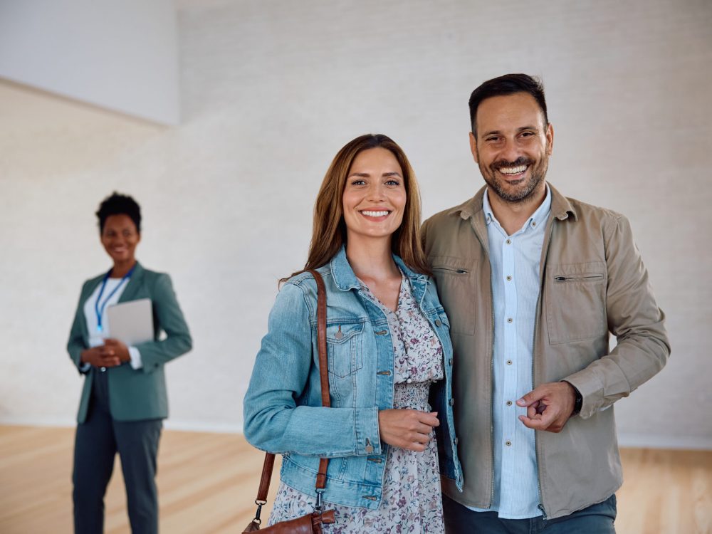 Happy couple buying a new house and looking at camera. Happy couple of real estate buyers looking at camera. Their agent is in the background.